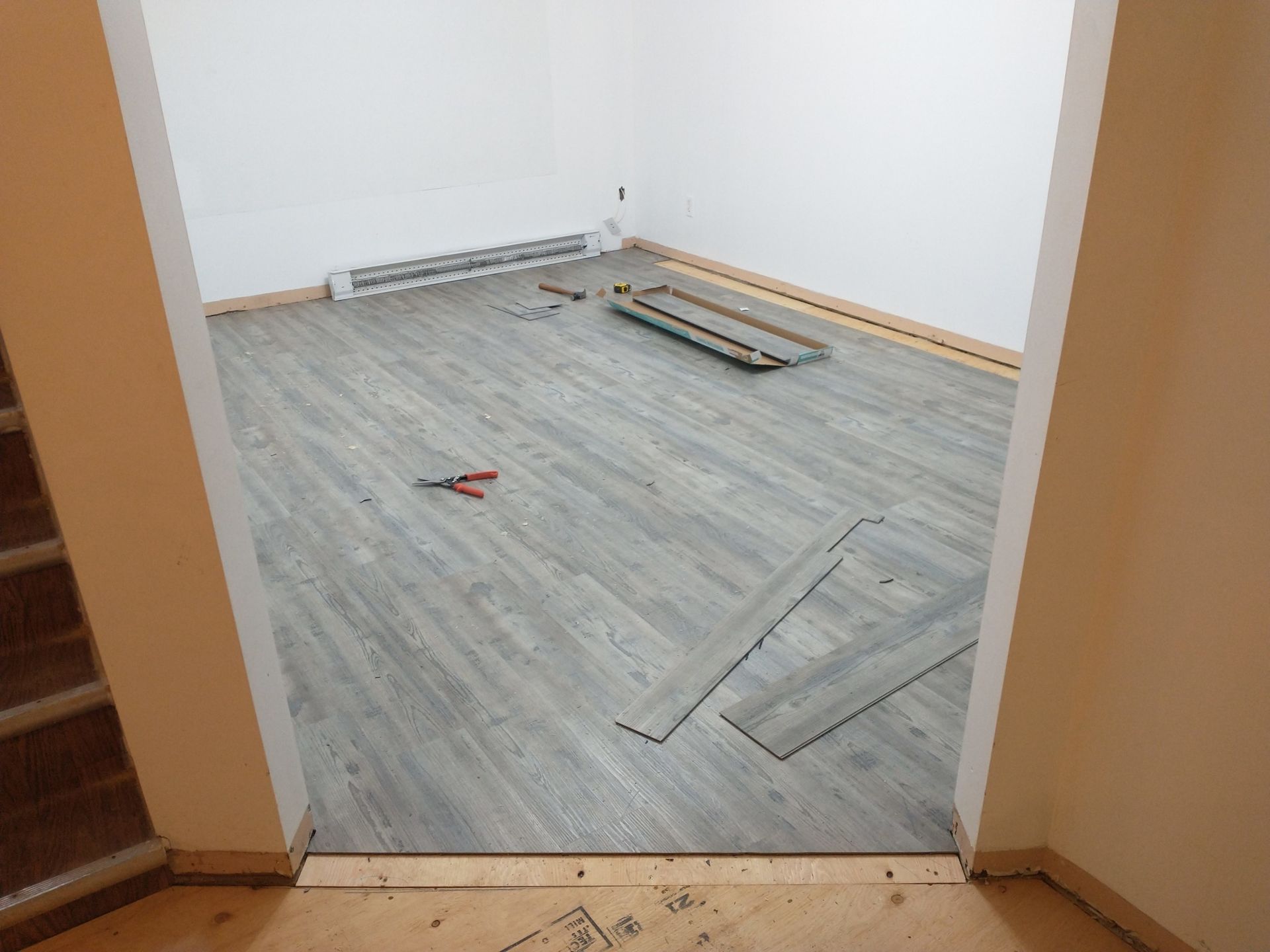 A room with partially installed light grey wood-look flooring, baseboard heating, and tools on the floor.