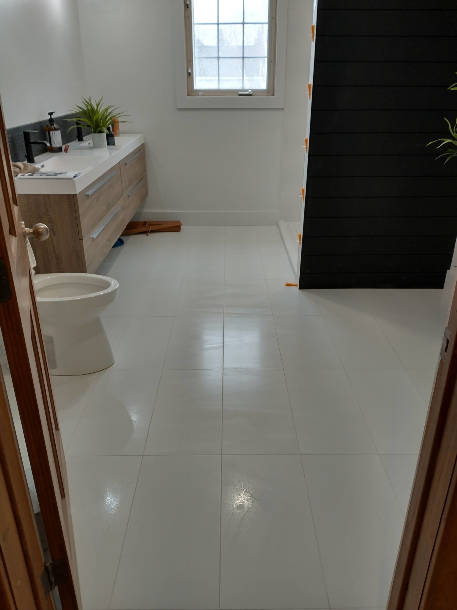 A bathroom with a floating light-wood vanity, white toilet, and glossy white floor tiles, next to a black-paneled wall.