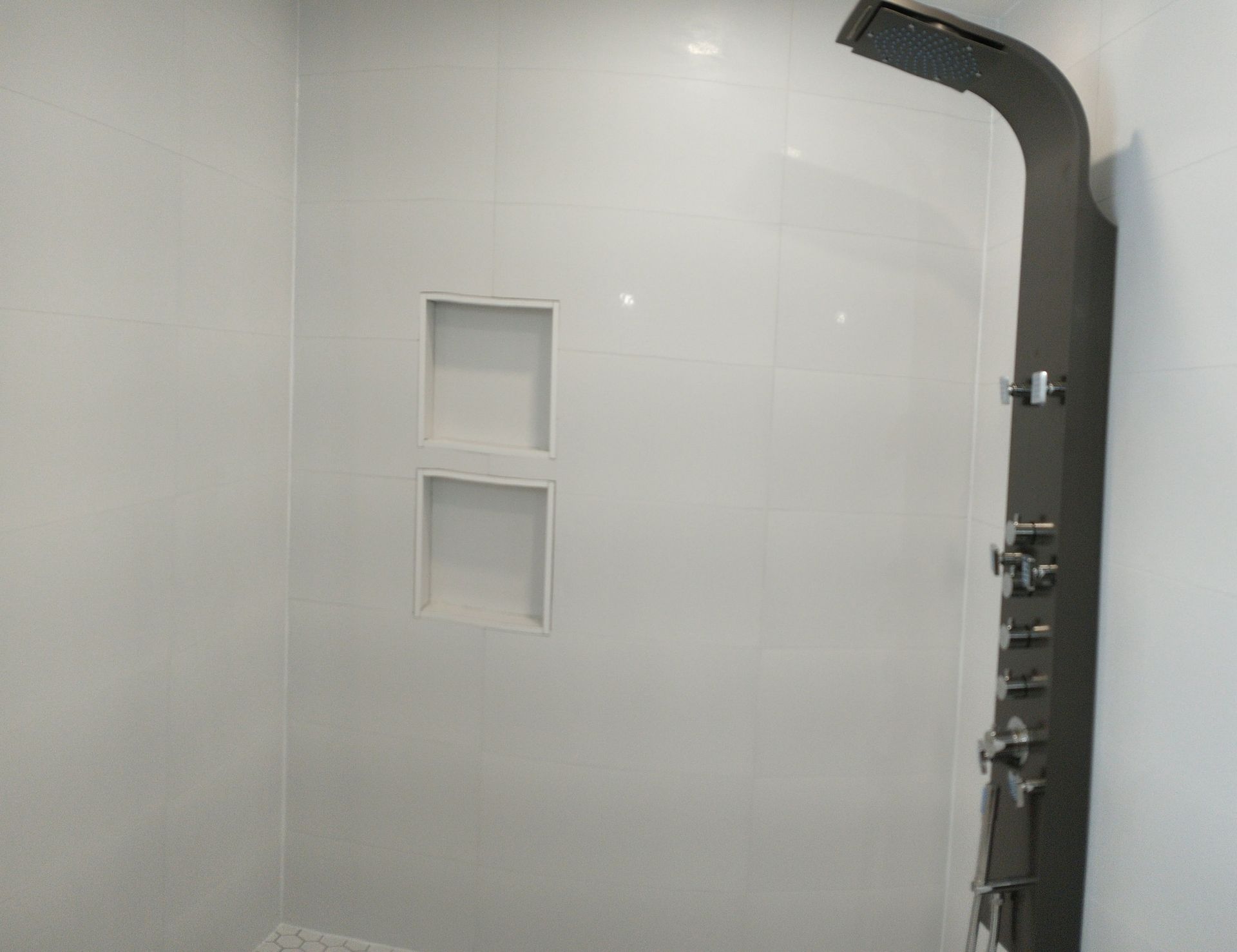 A modern shower stall featuring white tile walls, a built-in double-shelf niche, and a dark grey vertical shower panel.
