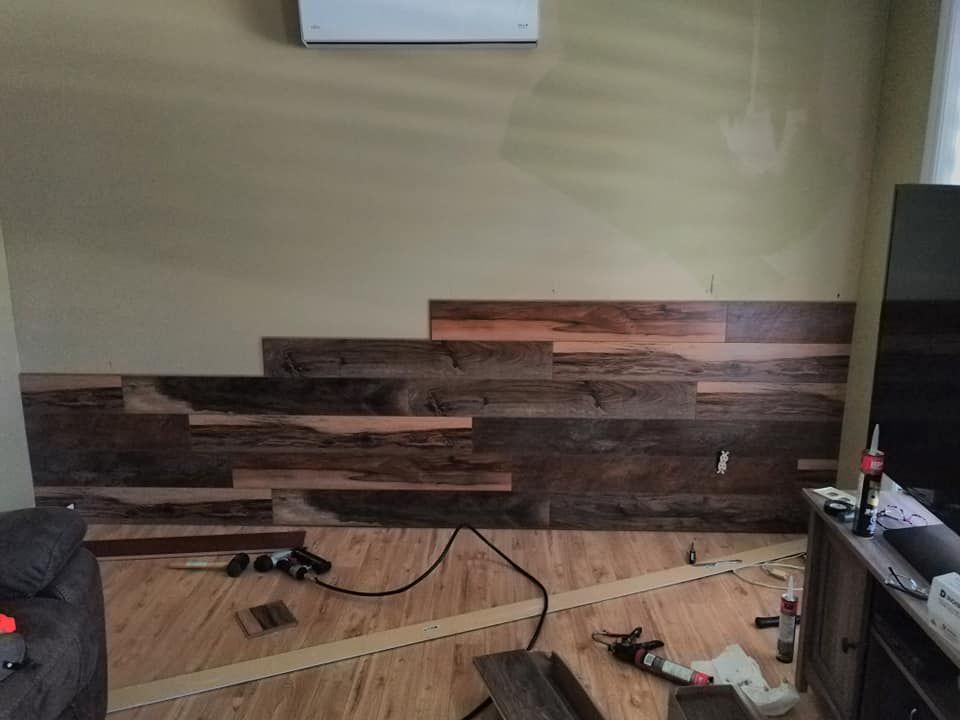 A living room wall with partially installed dark wood planks, with tools and construction materials on the floor nearby.