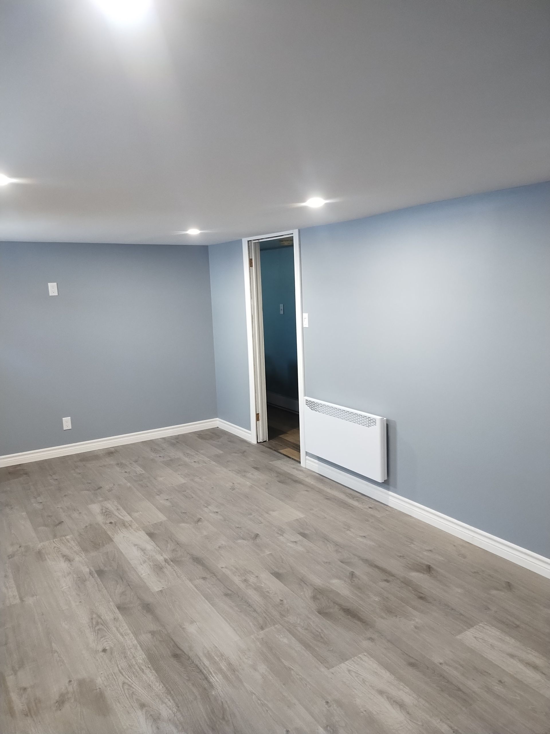 An empty room with gray walls, light wood-look flooring, recessed lighting, and a wall-mounted white heater.