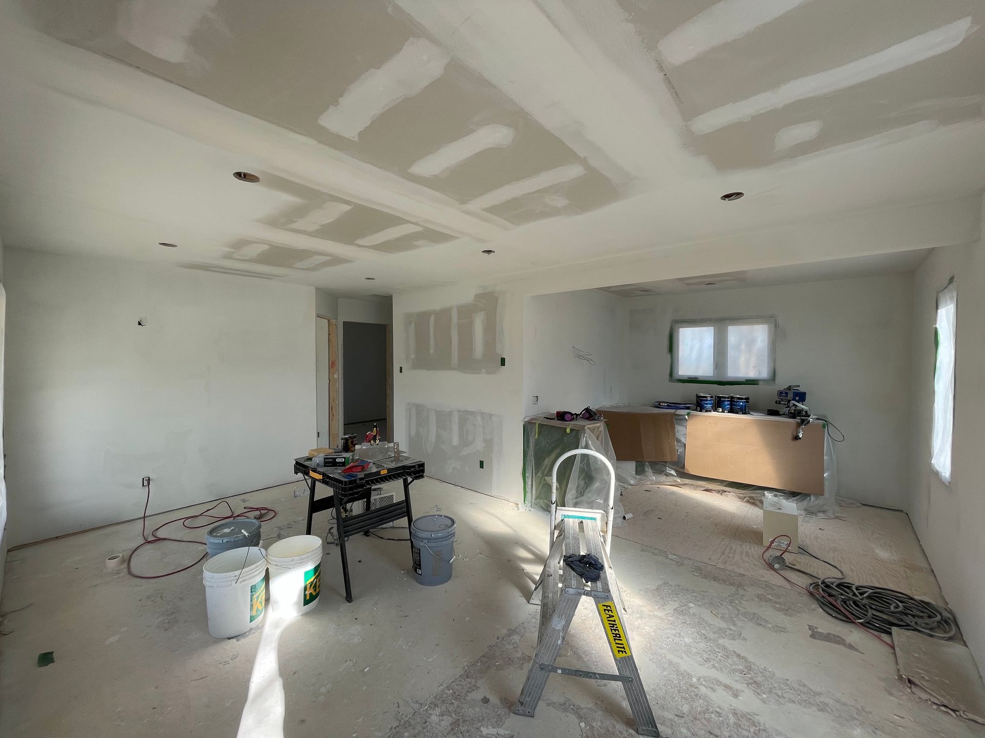 Interior of a house under renovation with drywall, construction tools, and a stepladder in an open floor plan room.