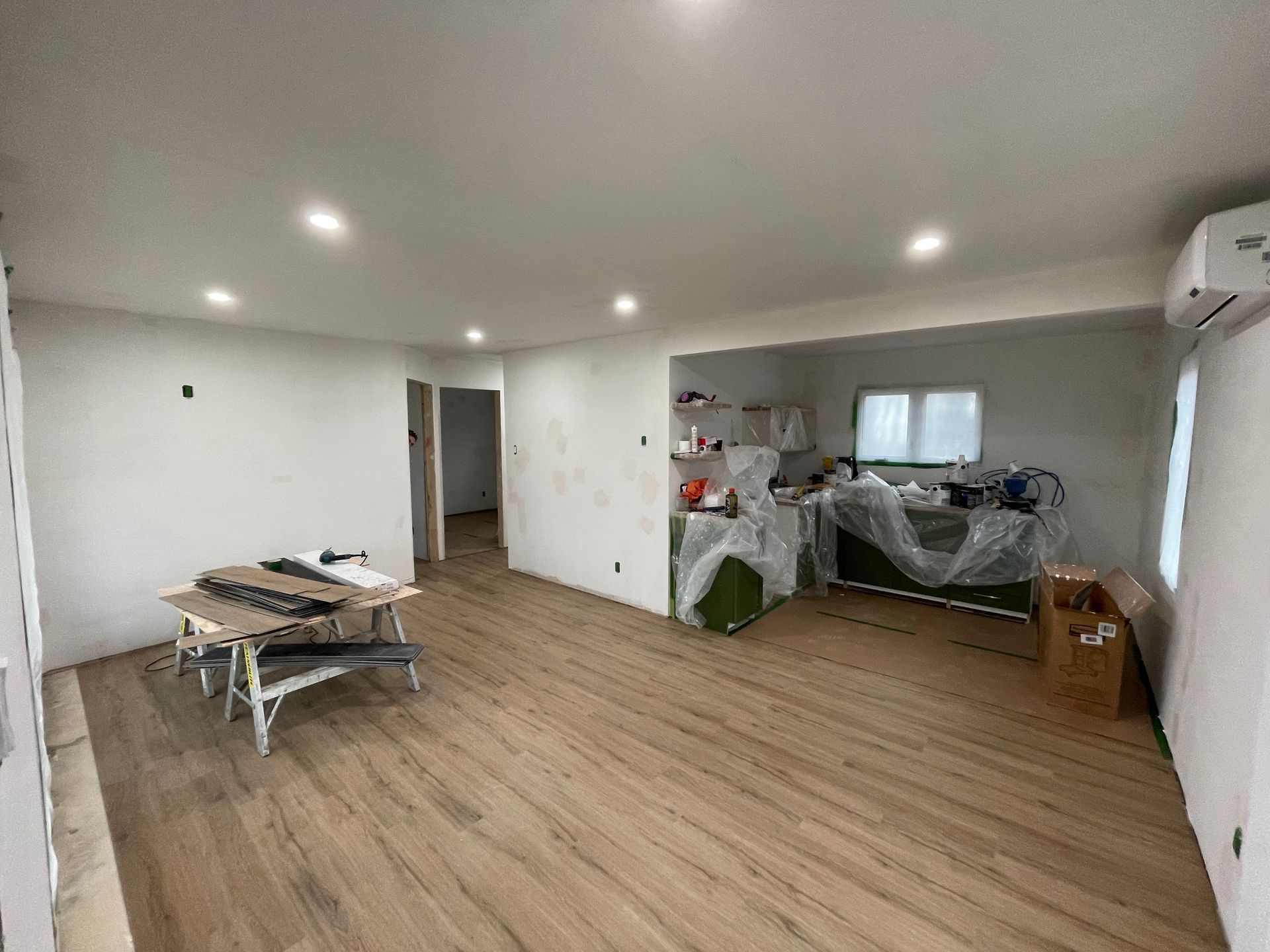 A wide view of a home interior under renovation with light wood flooring, white walls, recessed lighting, and equipment.