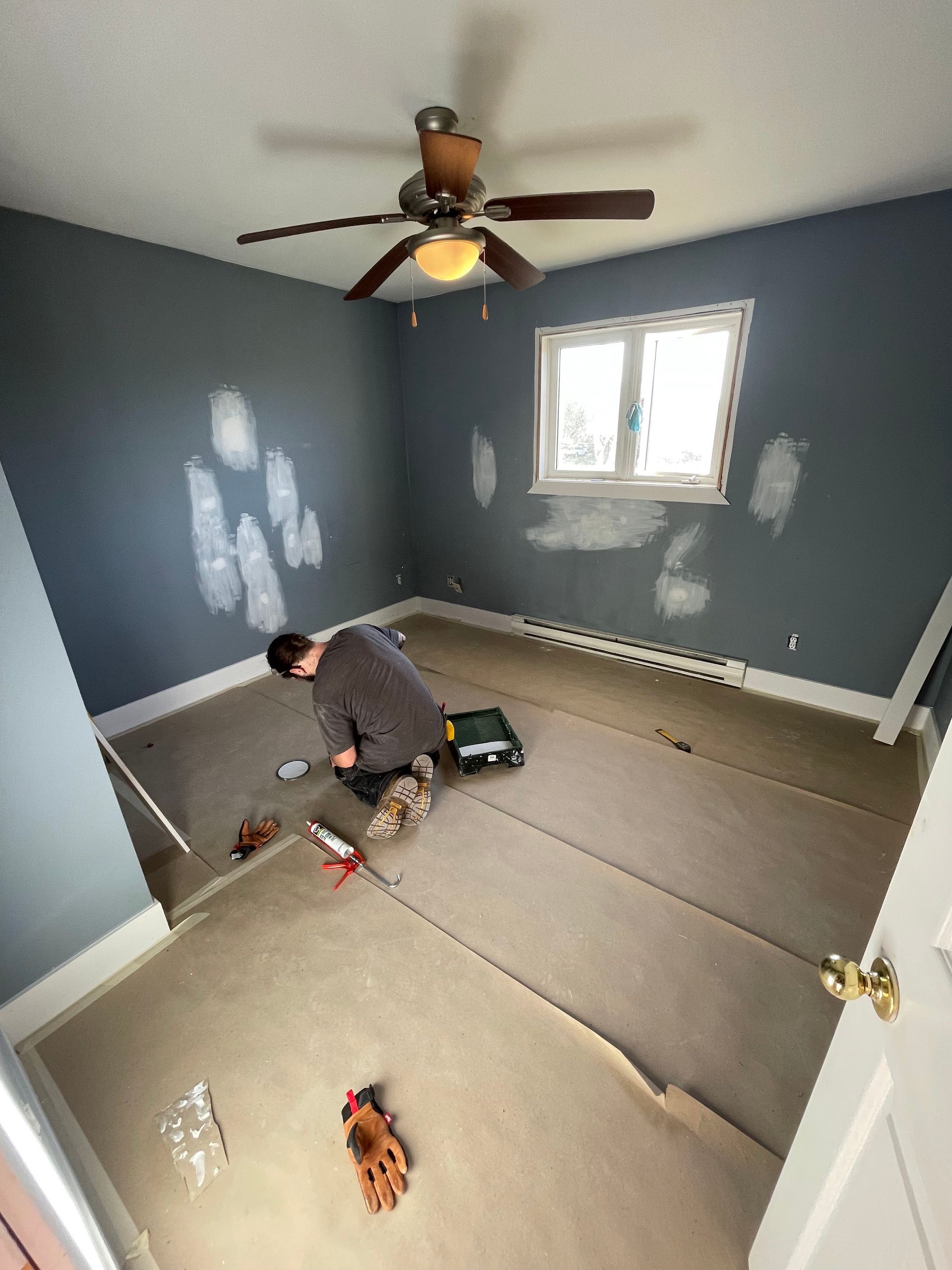A person works on flooring in a bedroom with gray walls and ceiling fan, with spackle patches visible on the walls.