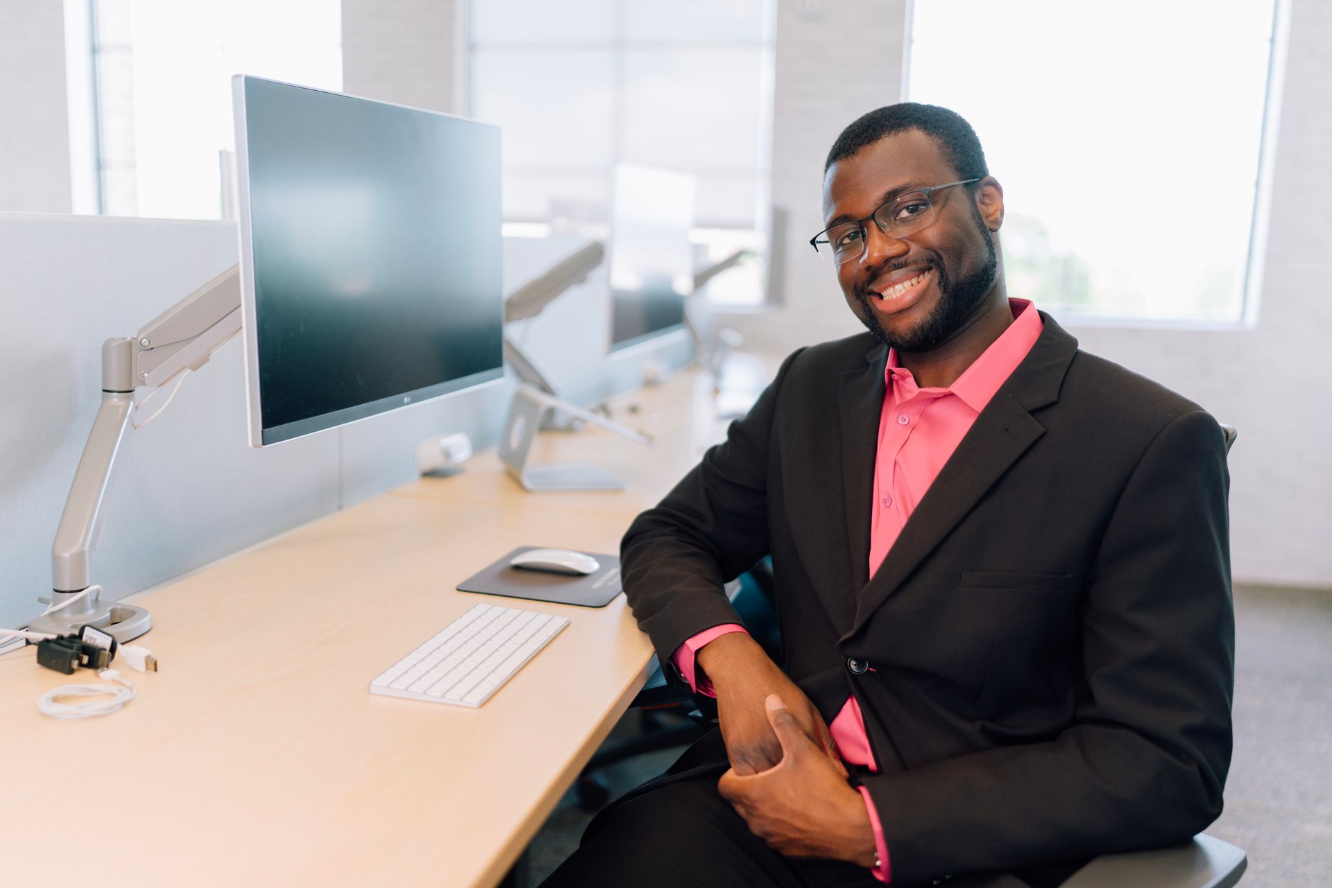 A man in a suit and pink shirt is sitting at a desk with a computer.