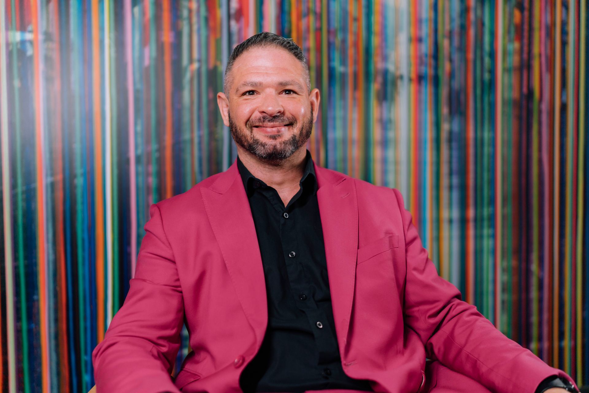 A man in a pink suit and black shirt is sitting in front of a colorful wall.