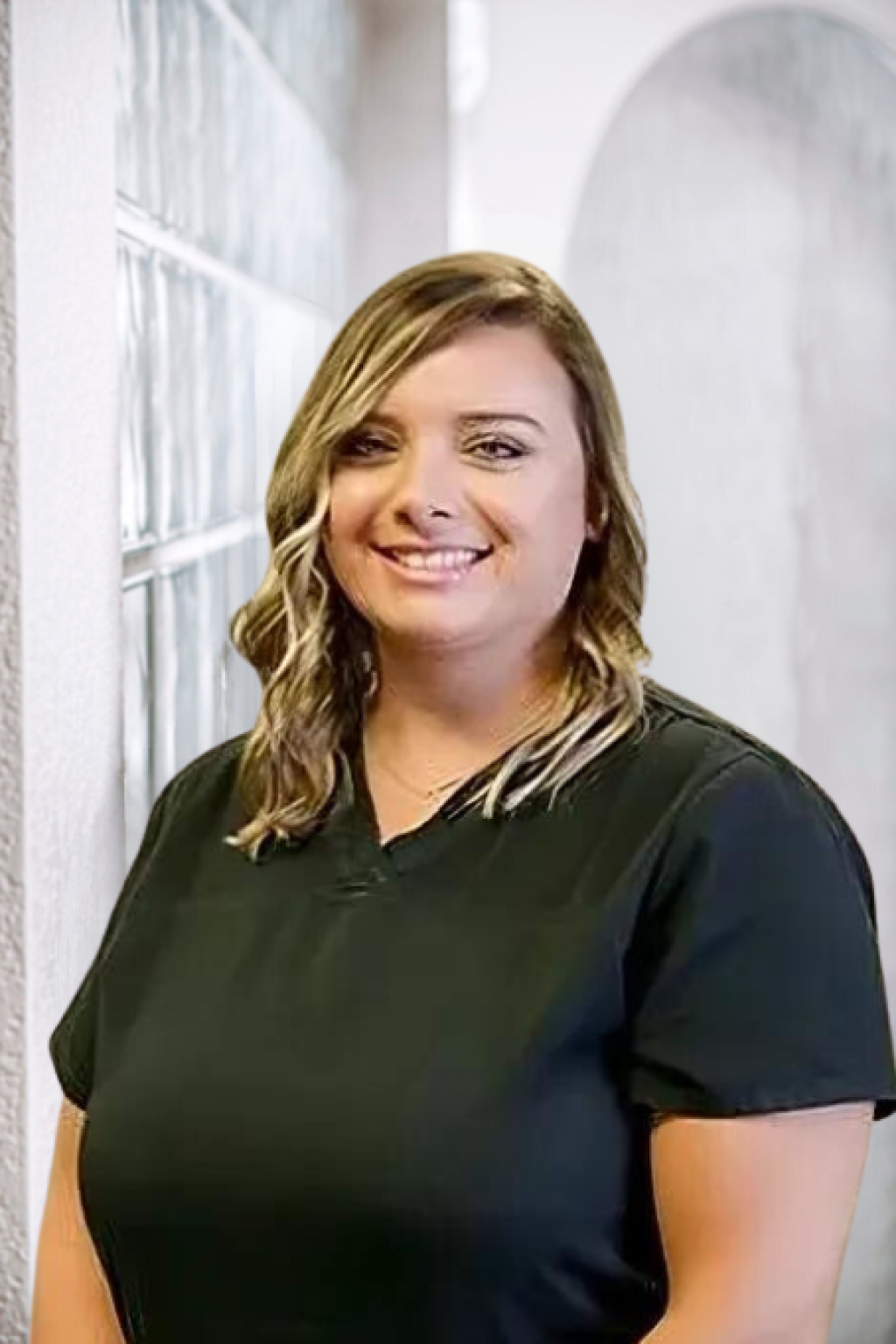 A woman in a black scrub top is smiling and standing in front of a window.