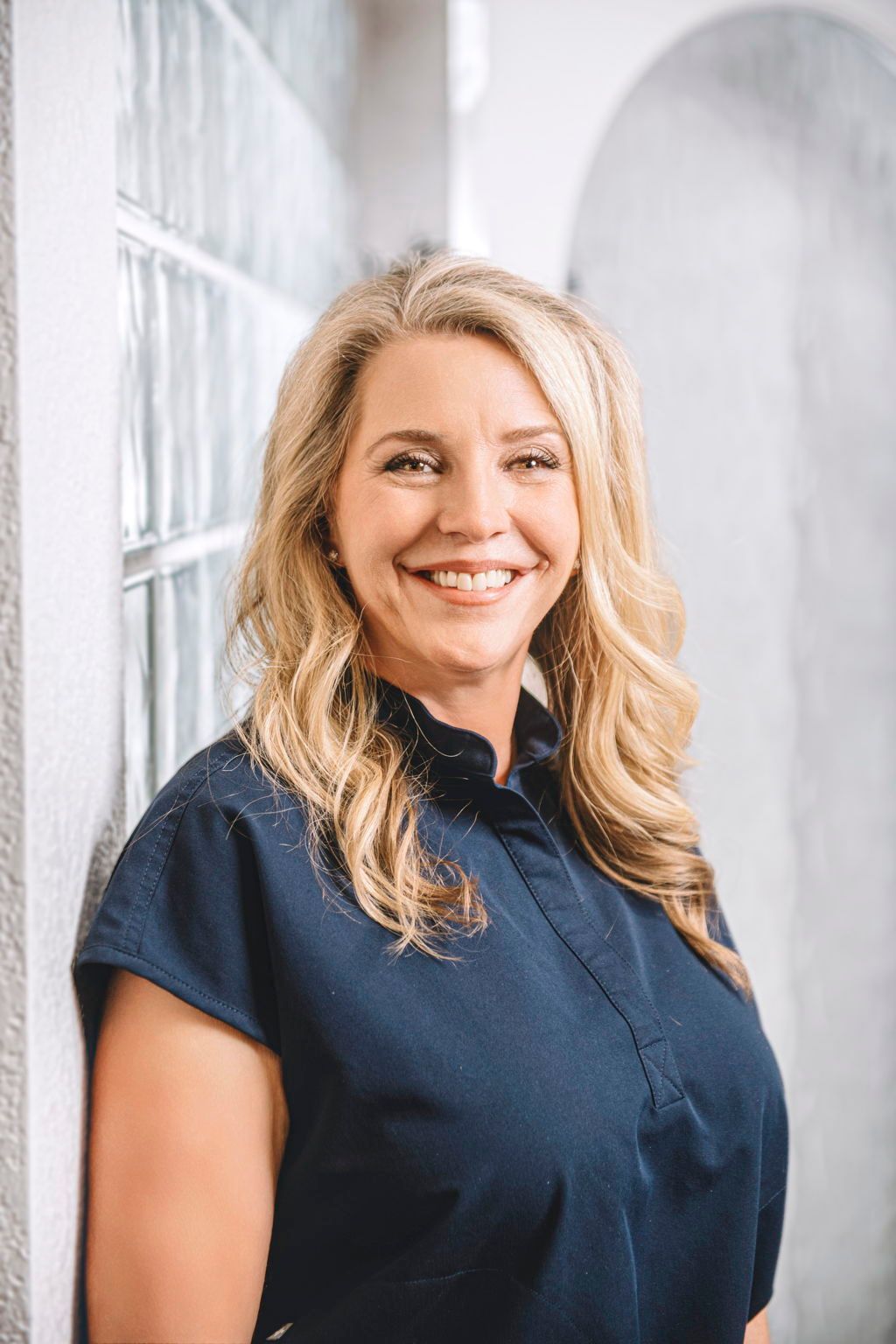 A woman in a blue shirt is leaning against a wall and smiling.