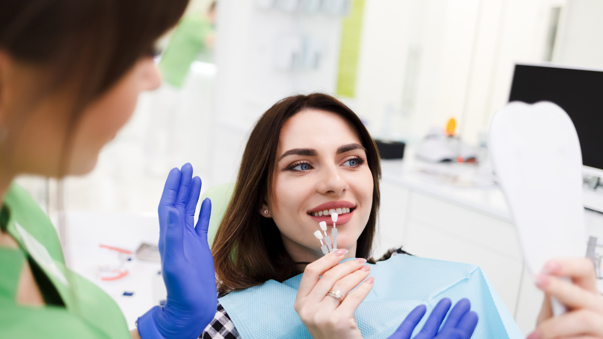 A woman is sitting in a dental chair looking at her teeth in a mirror.