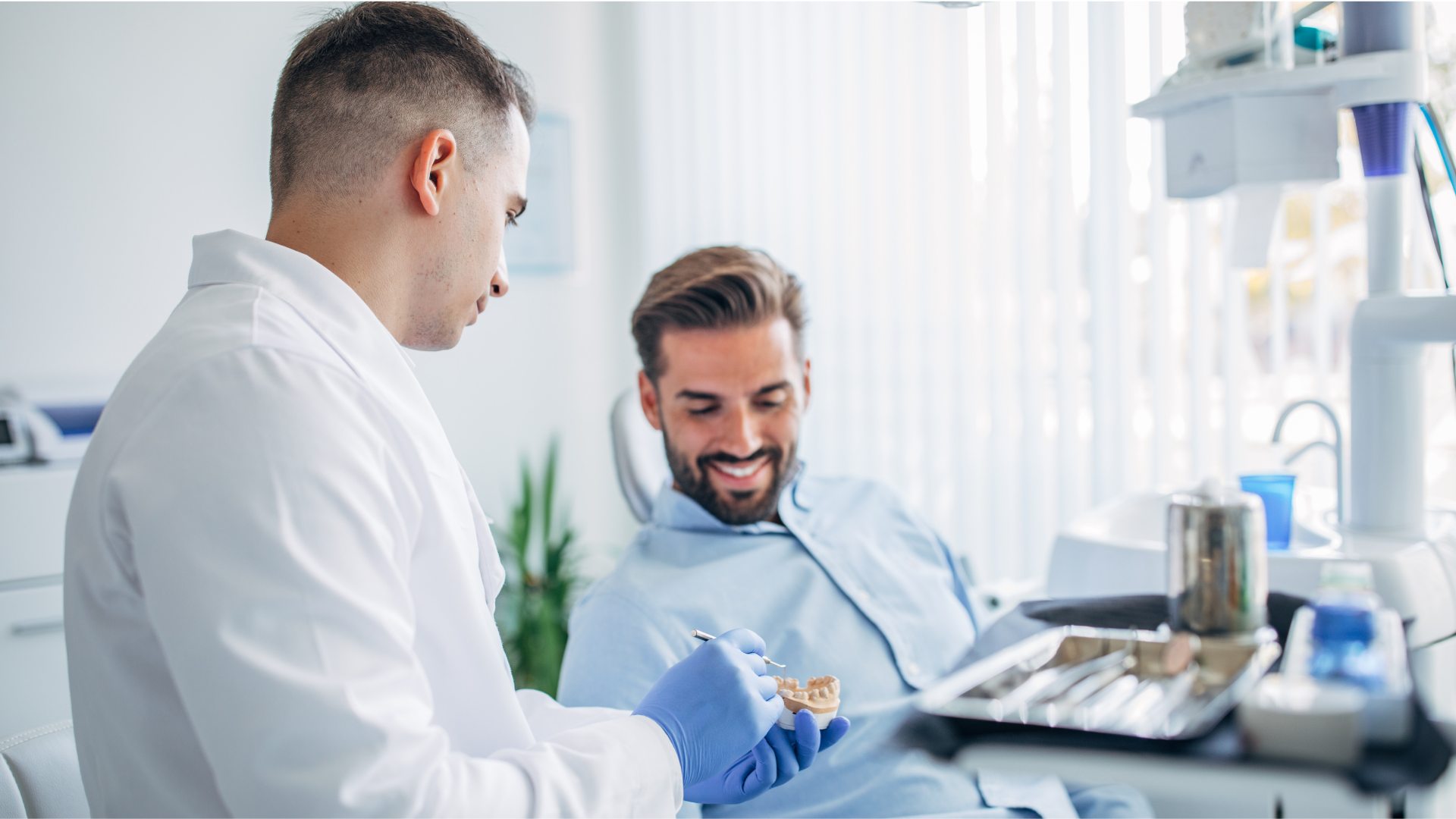 Dentist showing a patient a dental model in a brightly lit office. The patient smiles.