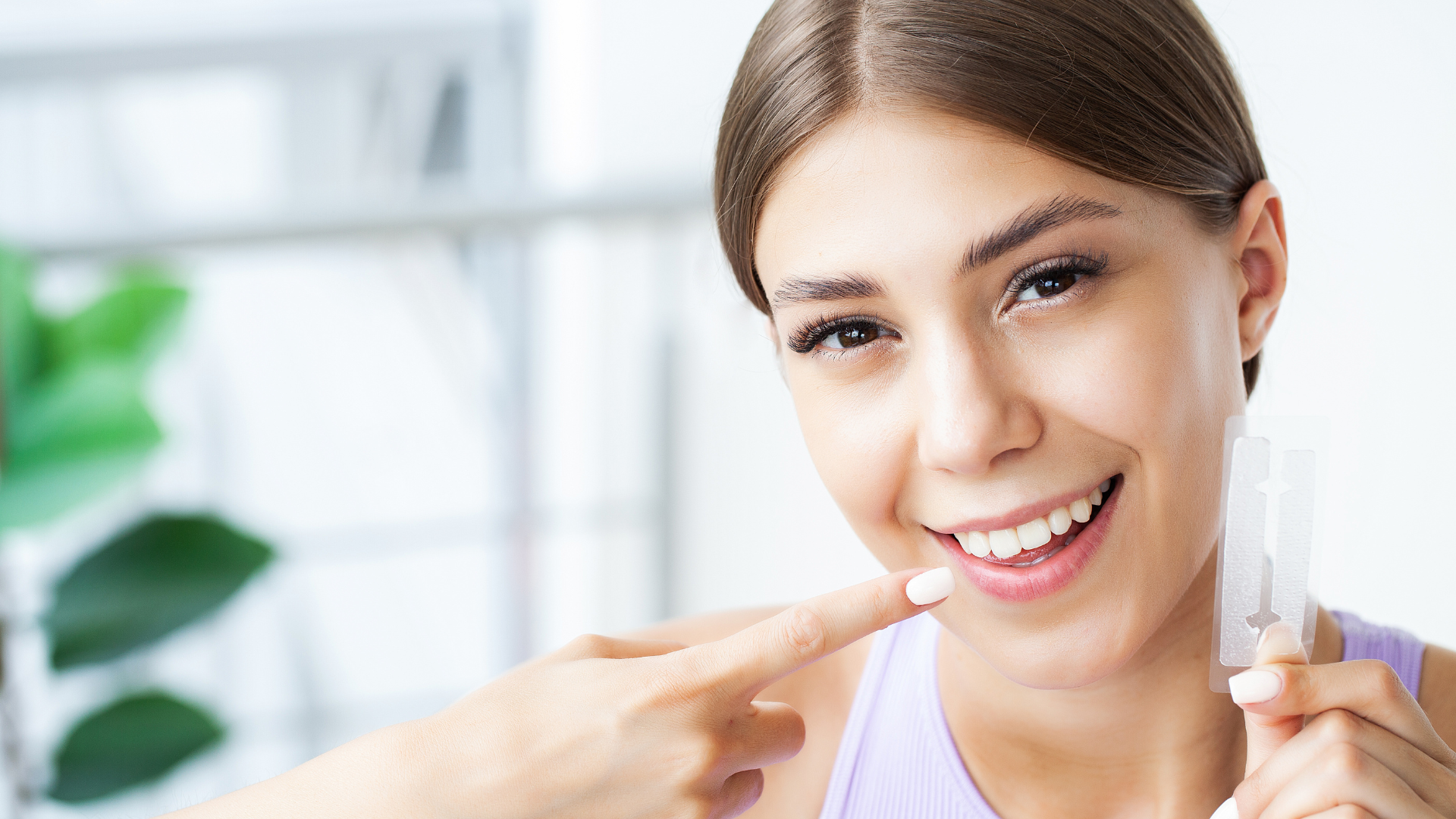 a woman is smiling and pointing at her teeth while holding a whitening strip .