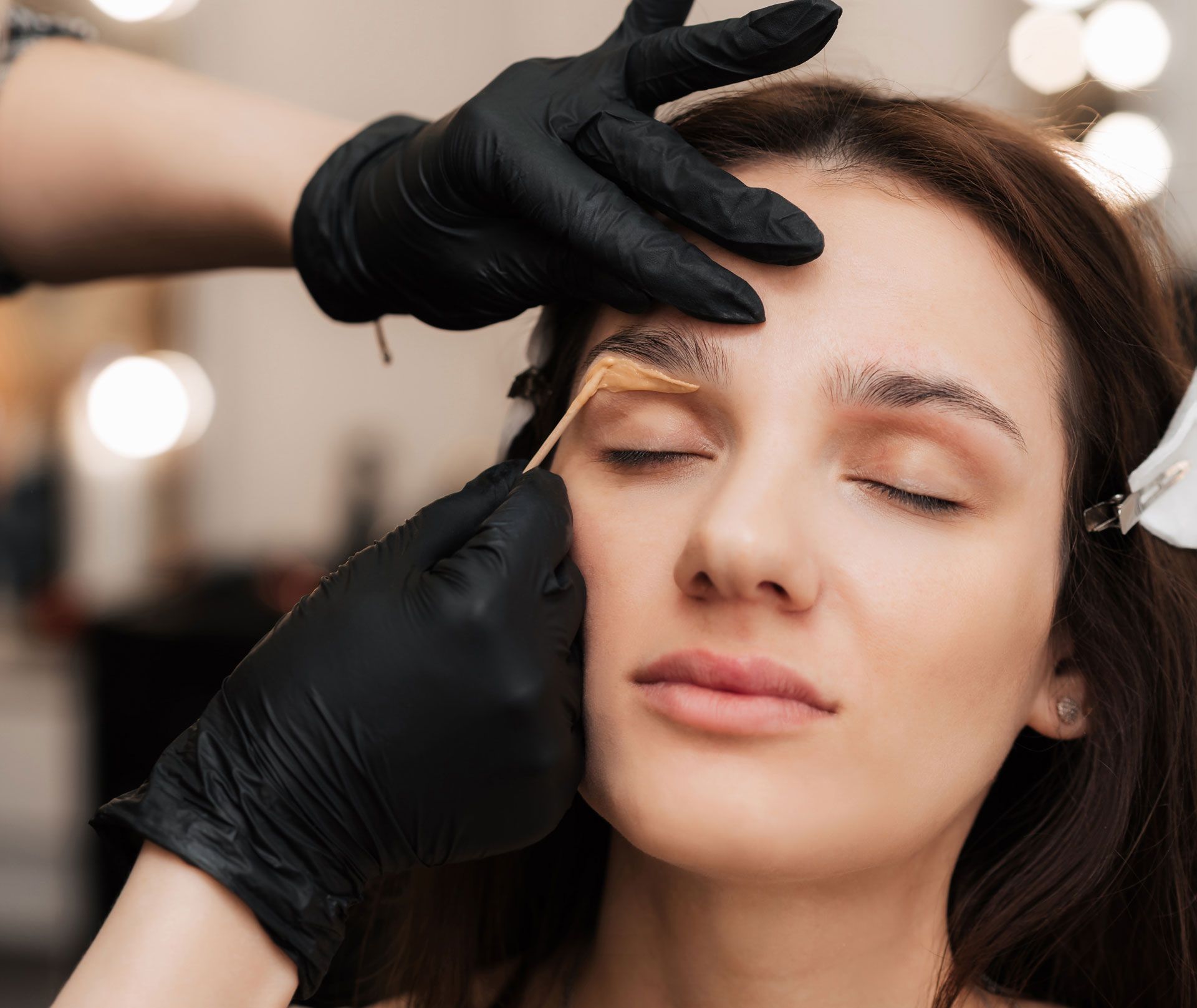 A beauty professional in black gloves applies wax to a client's eyebrow using a wooden spatula.