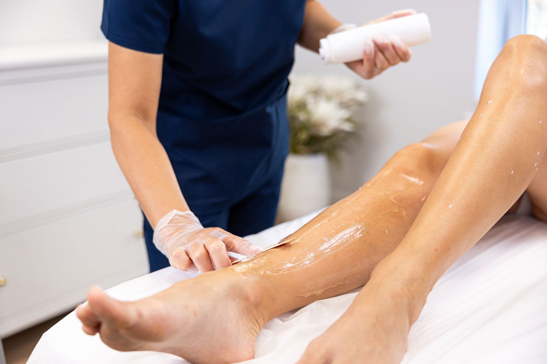 A person in scrubs and gloves applies hair removal wax to a client's leg at a clinic.