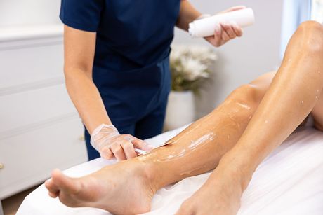 A person in scrubs and gloves applies hair removal wax to a client's leg at a clinic.