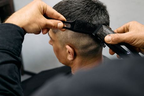 A barber uses electric clippers and a black comb to perform a precision fade on a person's hair.