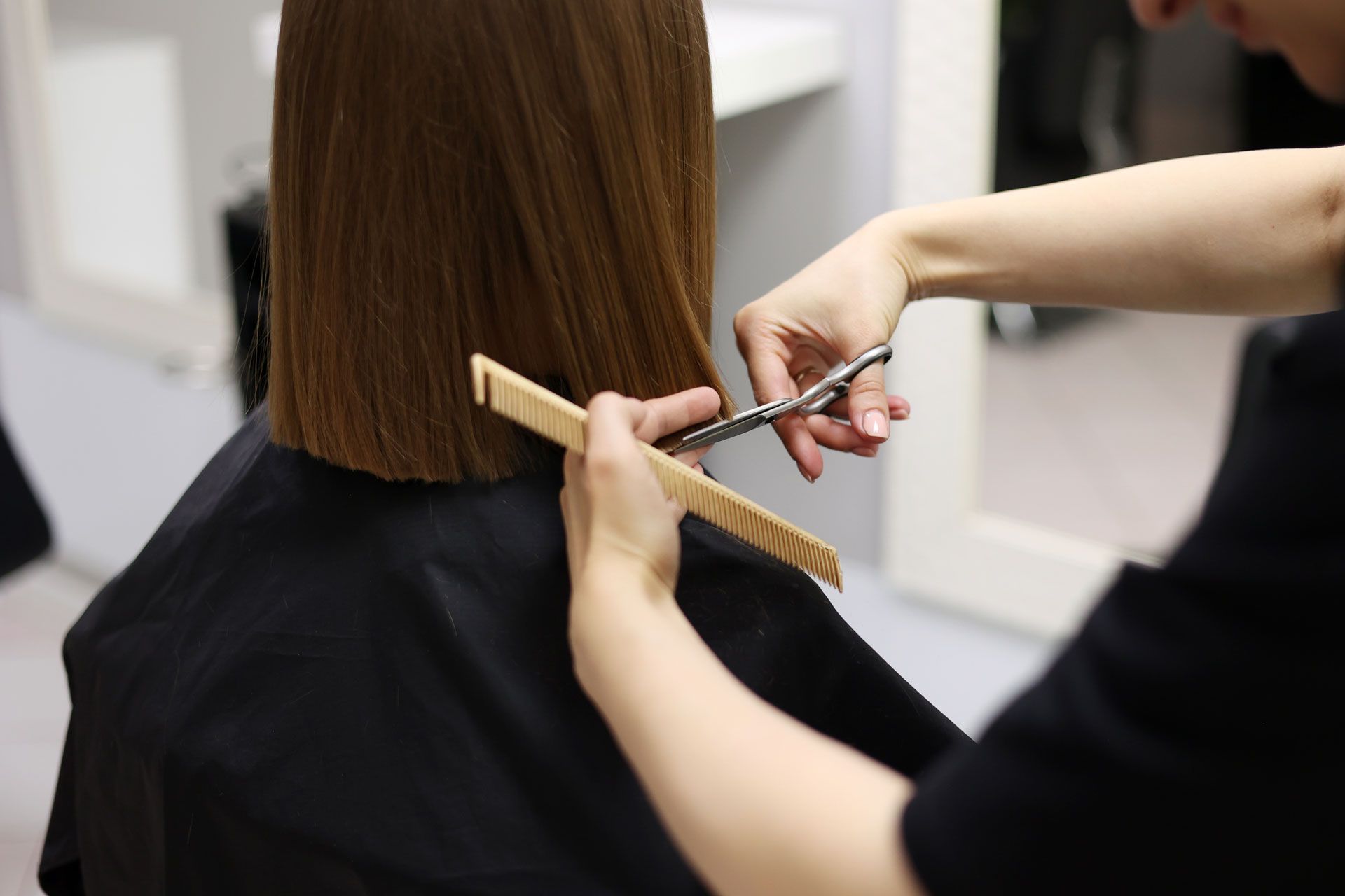 A stylist trims the straight, brown hair of a person wearing a black cape in a salon.
