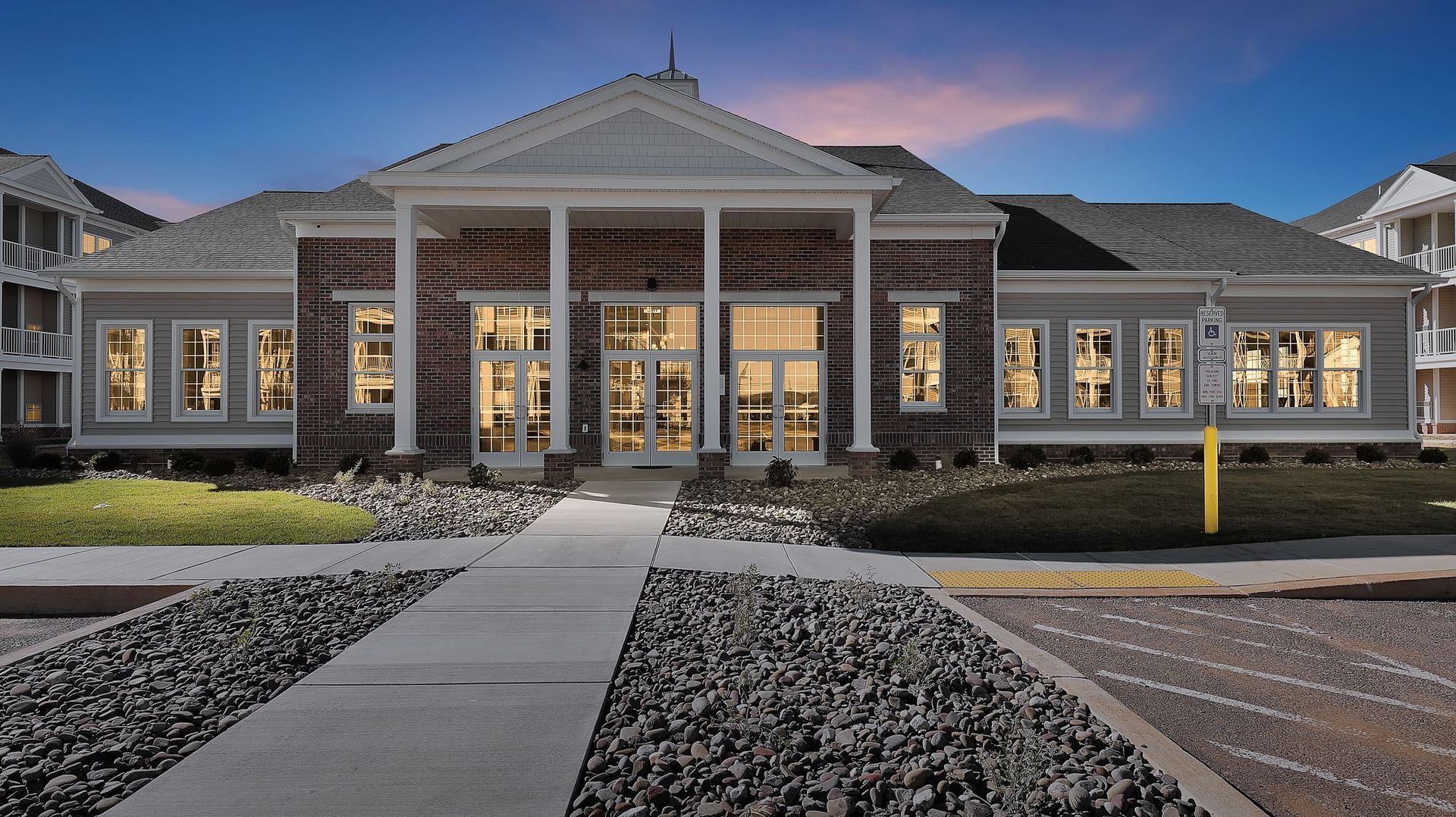 Apartment building entrance with columns, windows, and a walkway. Gravel and dusk lighting.