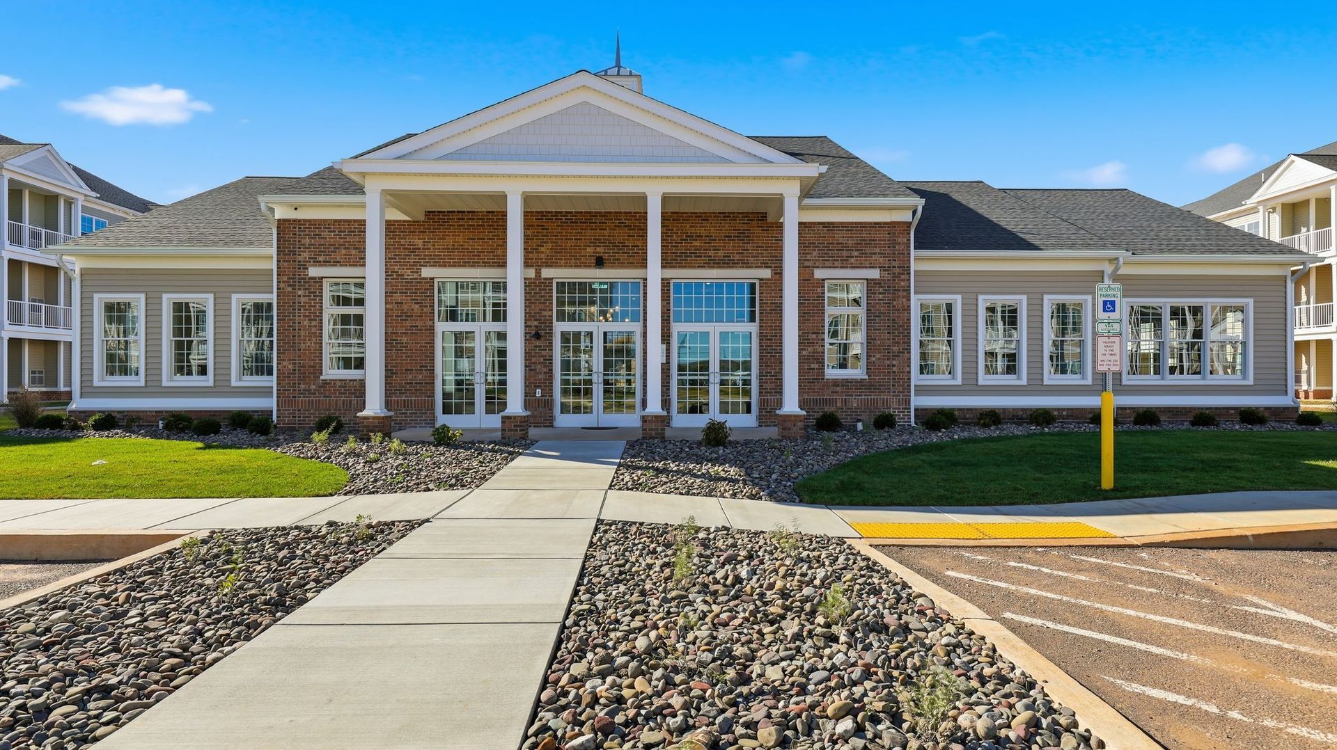 Apartment building entrance with brick facade, pillars, and sidewalk leading to glass doors, under blue sky.