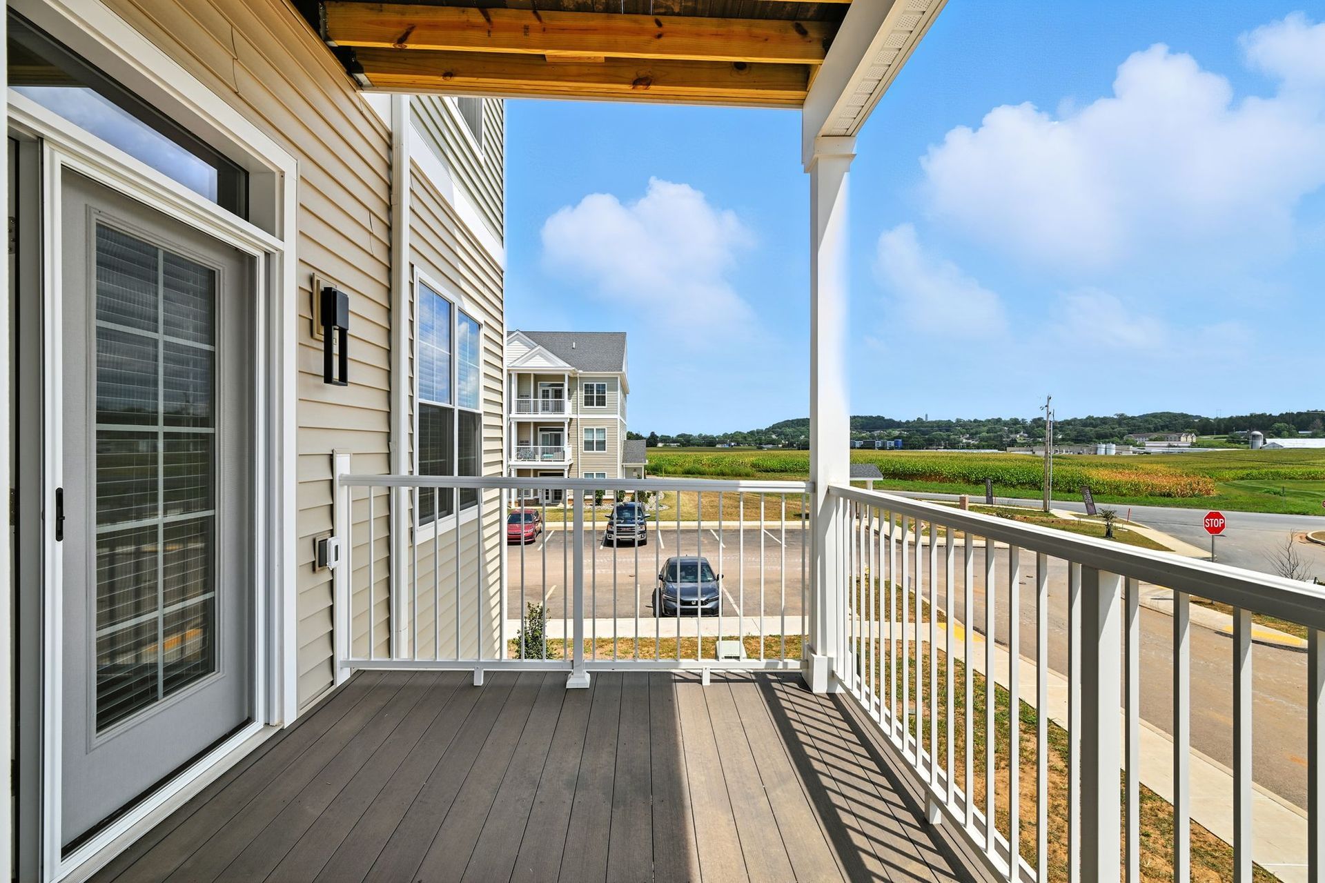 Balcony view overlooking parking lot and field, light tan siding, white railings, blue sky.