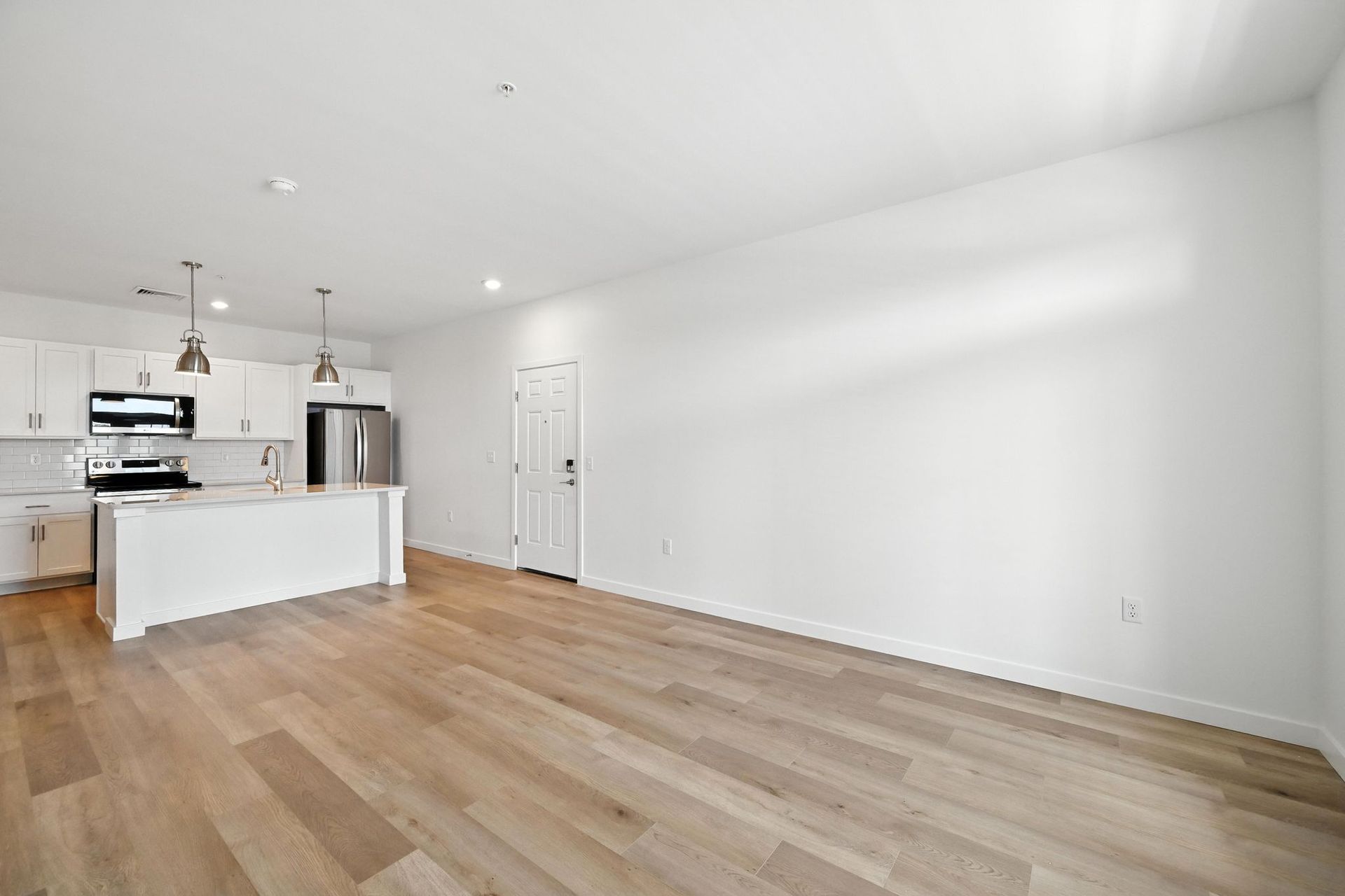 Empty open-concept living space with a white kitchen, stainless steel appliances, and wood flooring.
