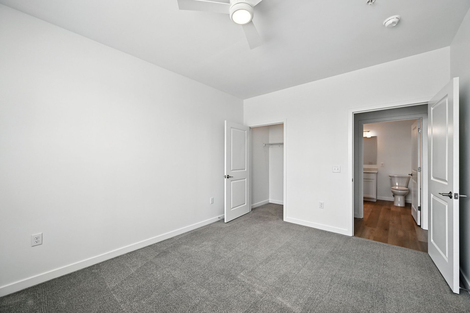 Empty bedroom with gray carpet, white walls, closet, and open doorway to bathroom.