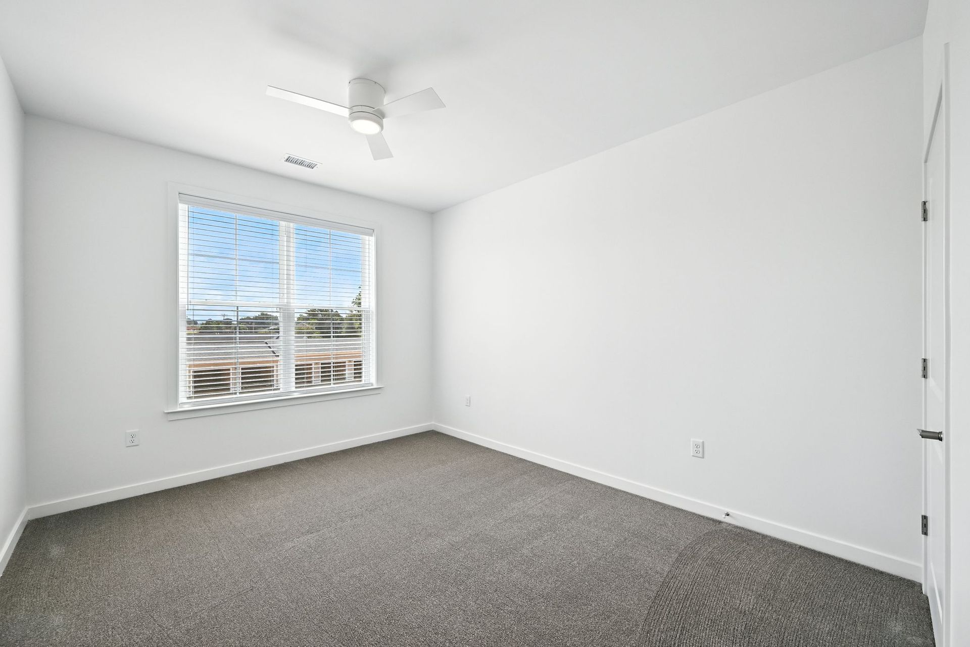 Empty bedroom with gray carpet, a window, and a ceiling fan. Walls are white.