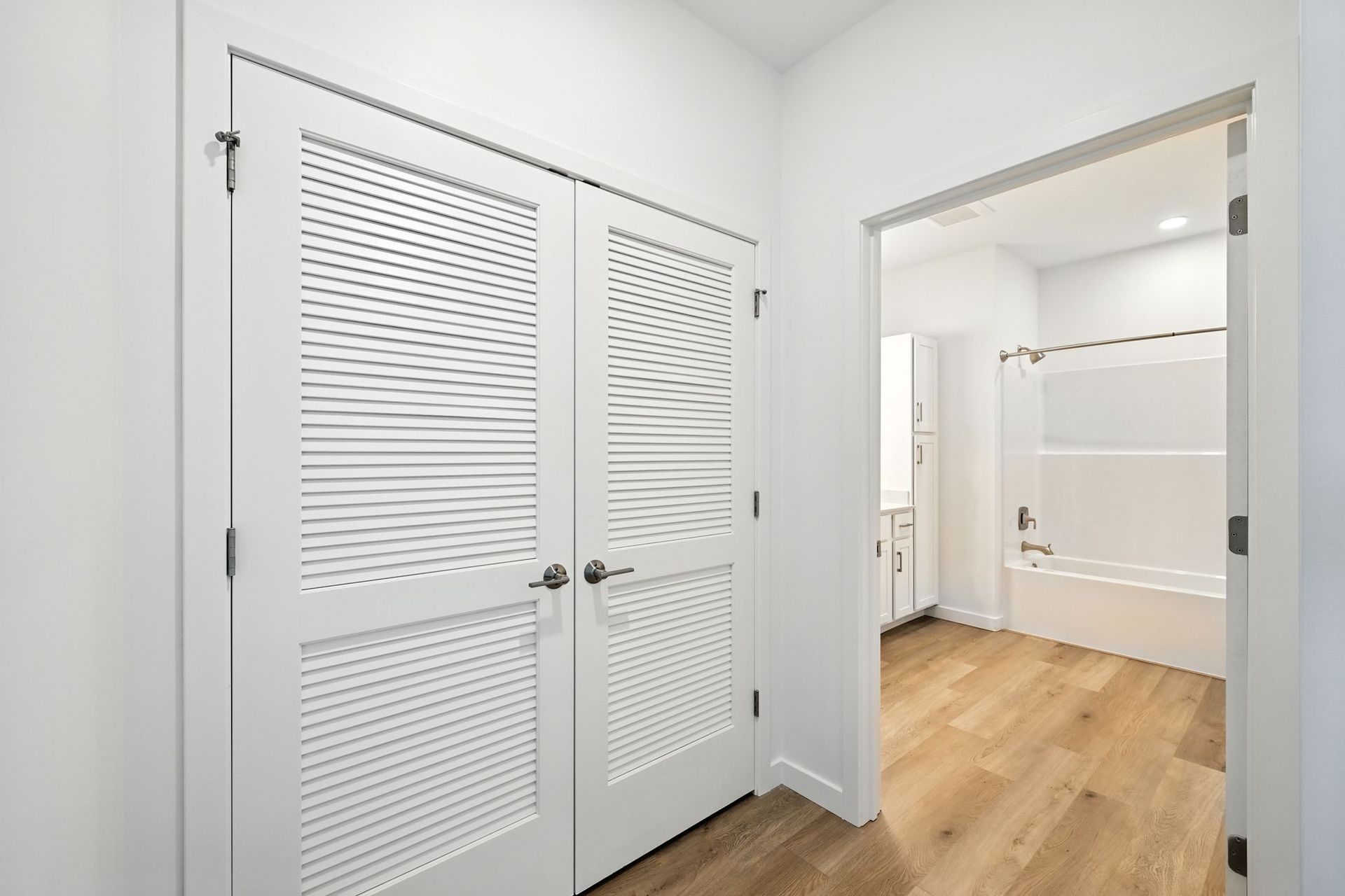 White hallway with louvered doors on the left, open doorway to a bathroom on the right, light wood floors.