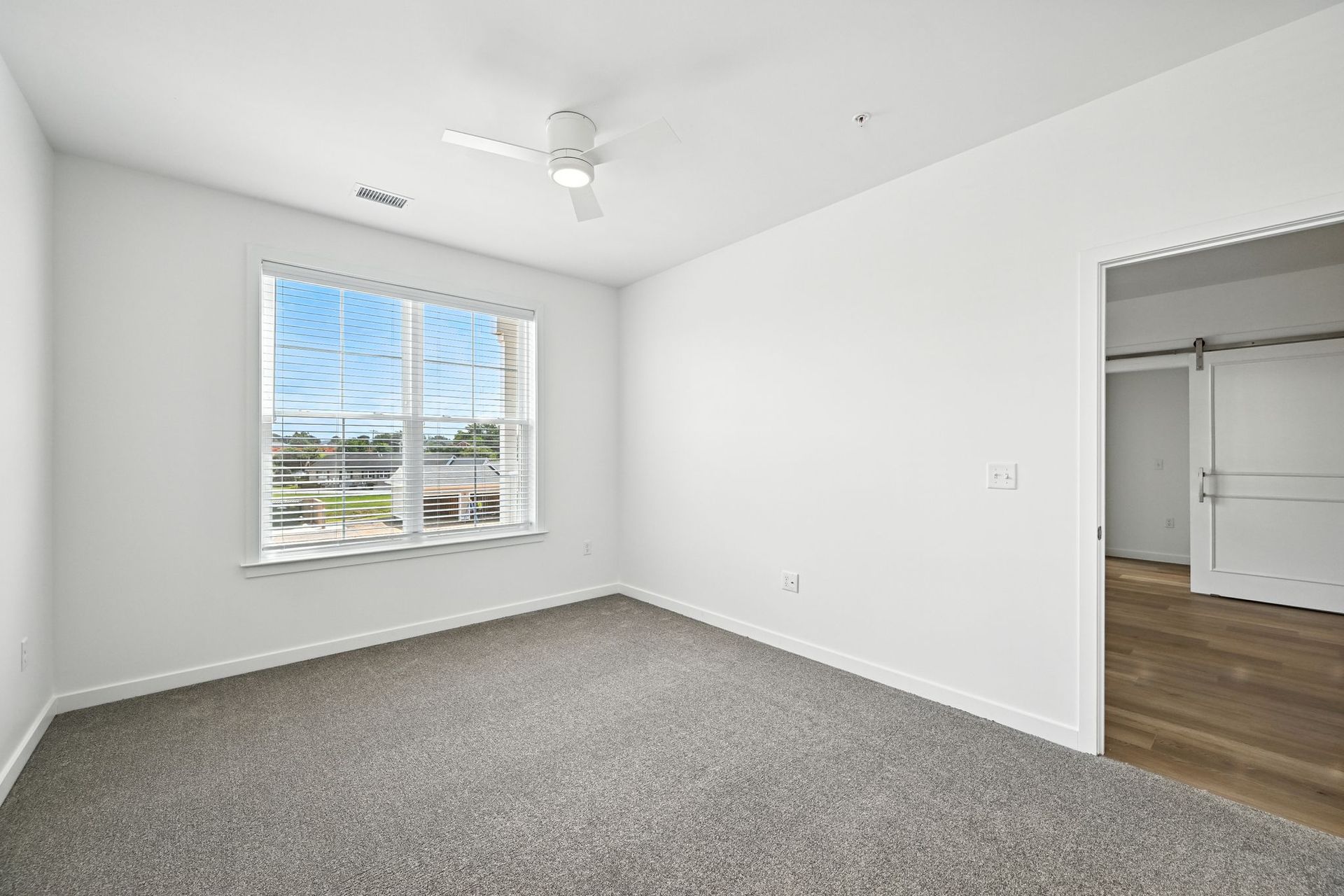 Empty bedroom with gray carpet, white walls, window, and open doorway to another room.