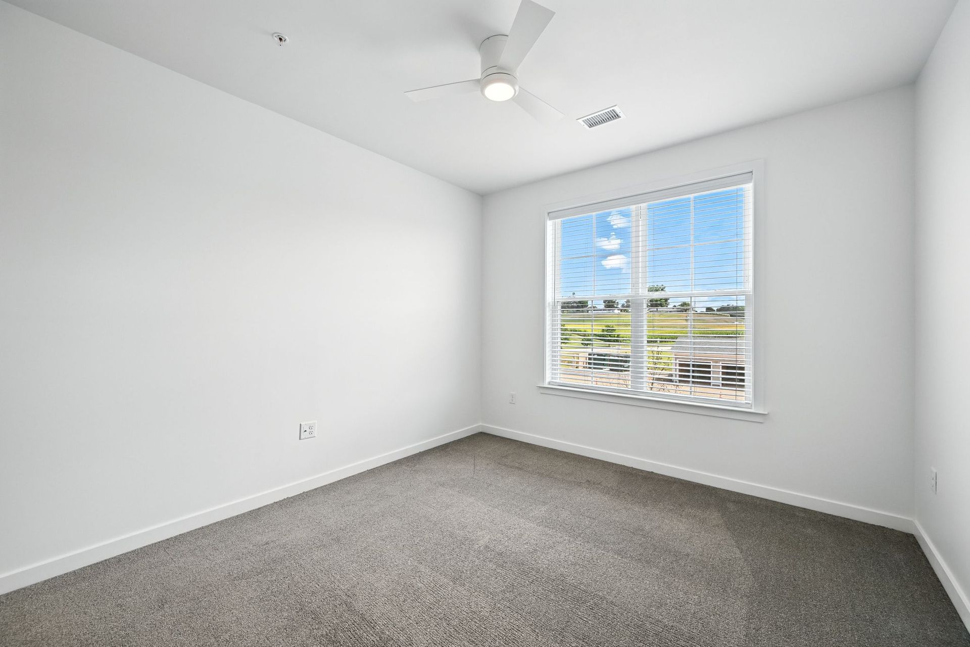Empty room with white walls, gray carpet, and a window with a view of greenery. Ceiling fan.