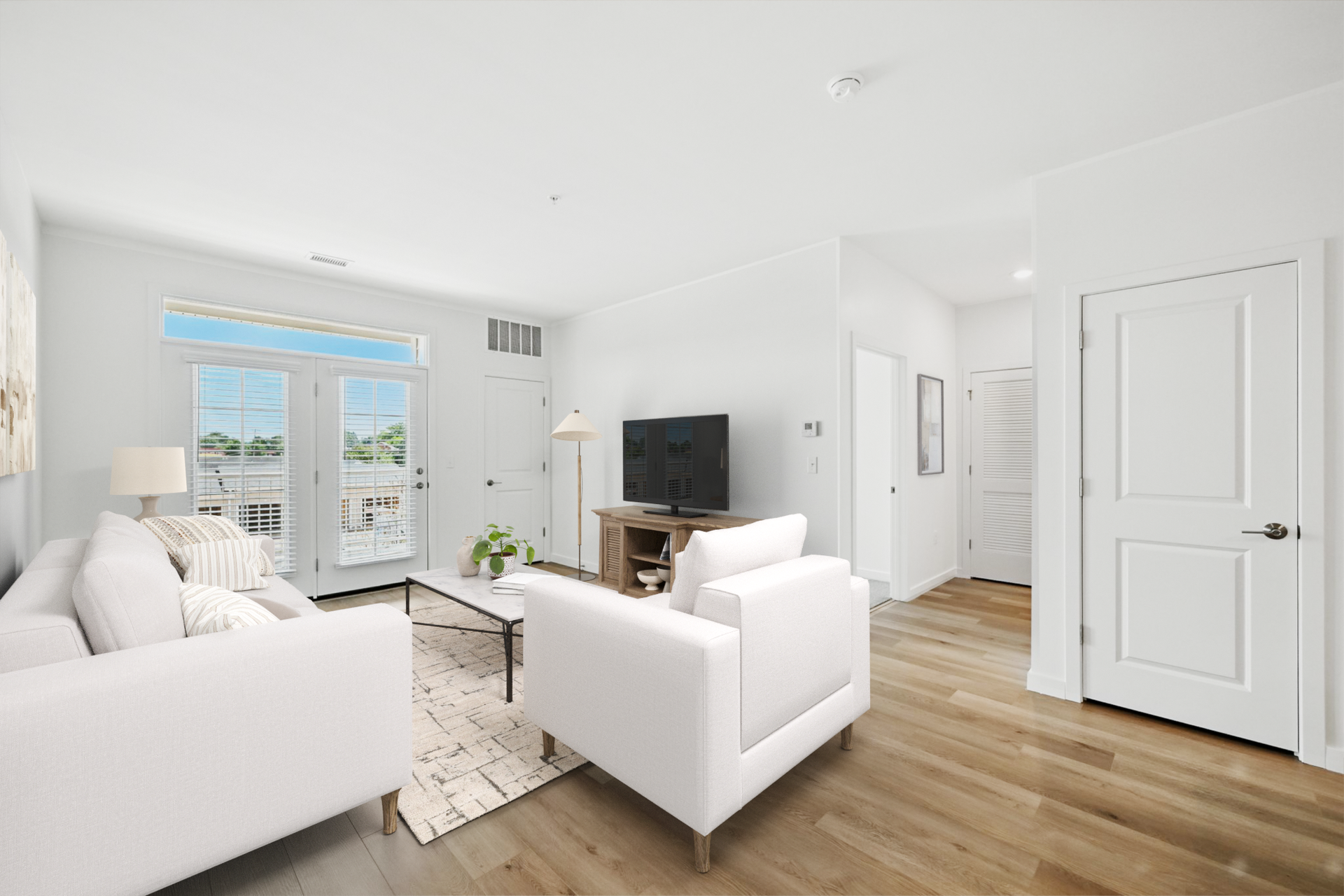 Bright living room with white furniture, wooden floors, and a TV. French doors lead to a balcony.