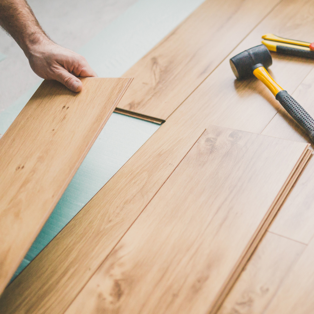 A hand installing light-colored wood laminate flooring with a rubber mallet and utility knife nearby.