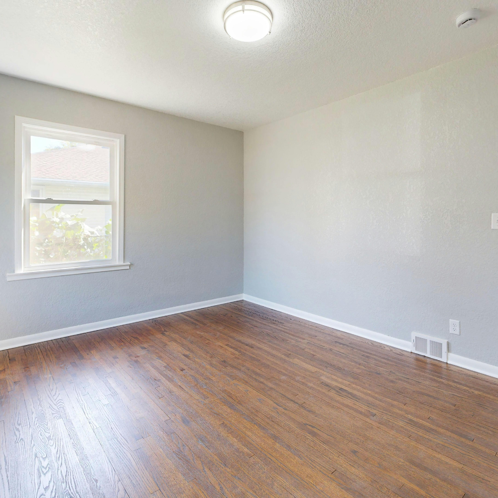 A bright, empty room with light gray walls, wooden flooring, a single window, and a ceiling light fixture.