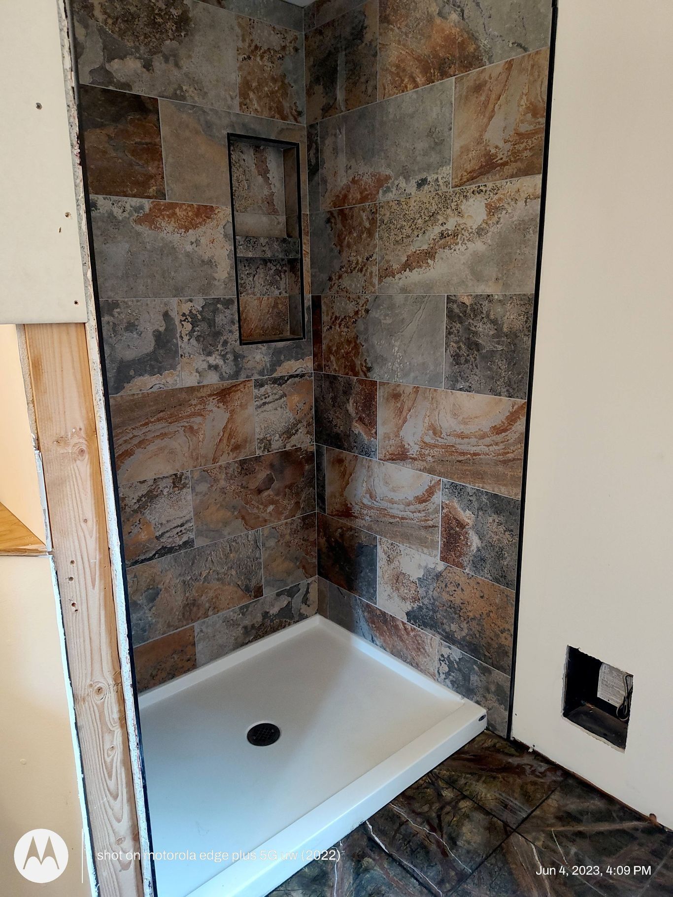 A partially finished shower stall with multicolored, textured stone tiles and a white base, currently under construction.
