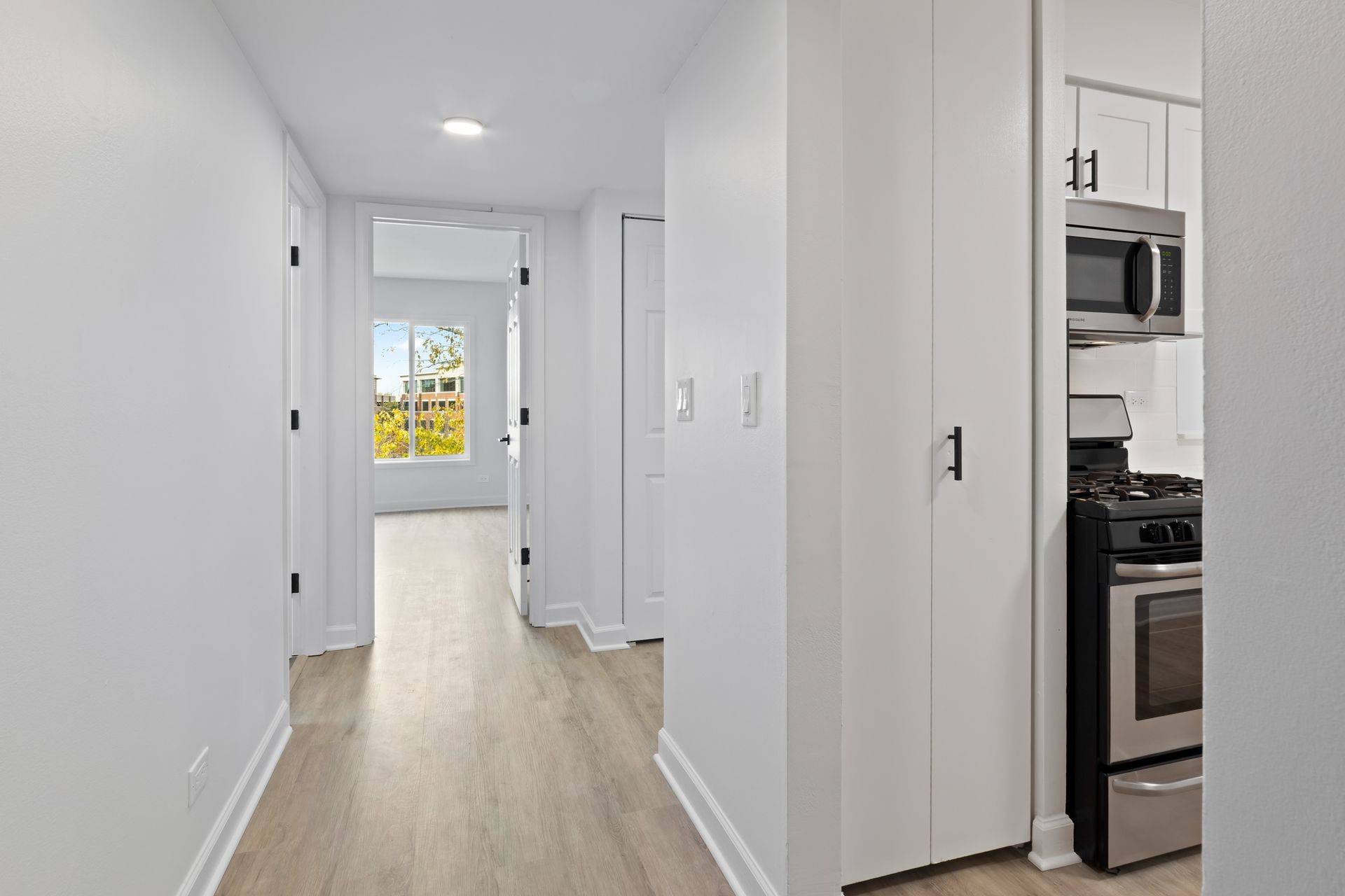 Hallway with light wood floor, white walls and doors, leading to a window. Kitchen is on right.