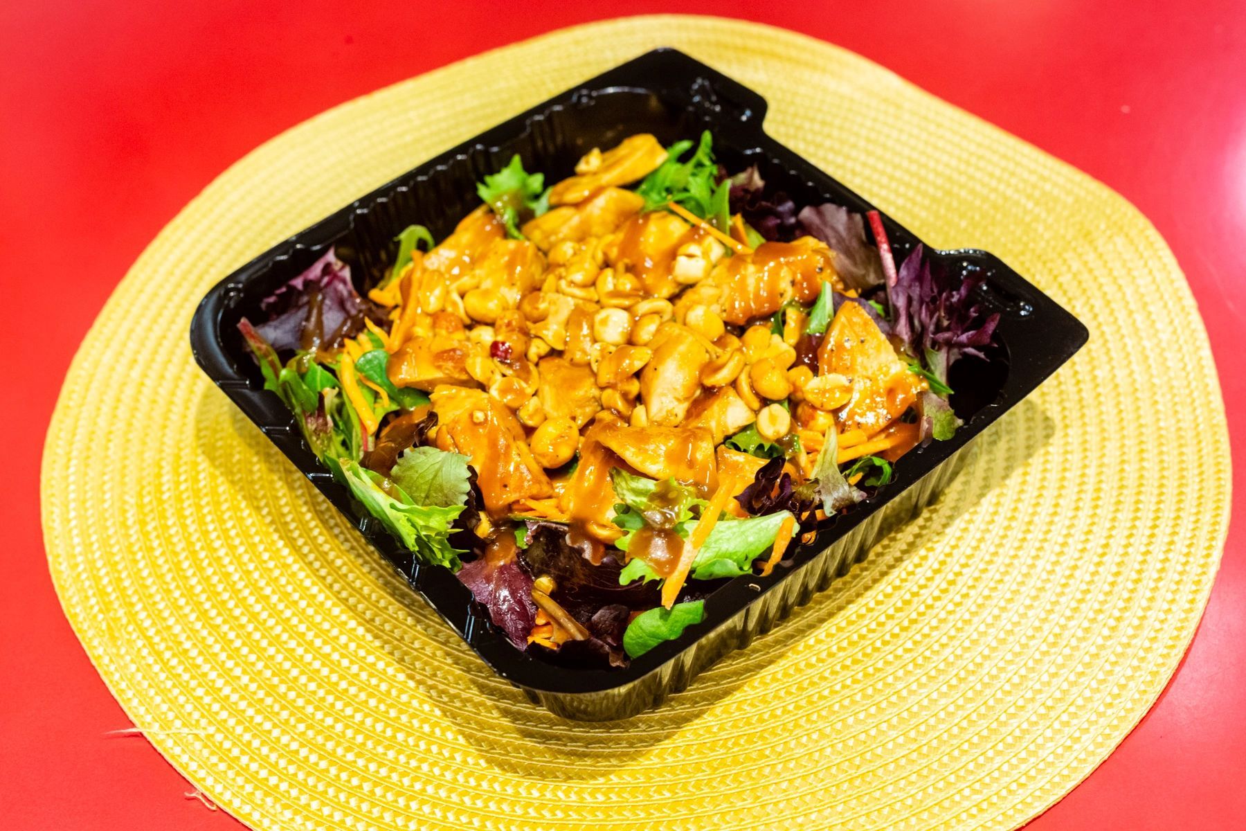 A salad in a plastic container is sitting on a yellow place mat on a table.