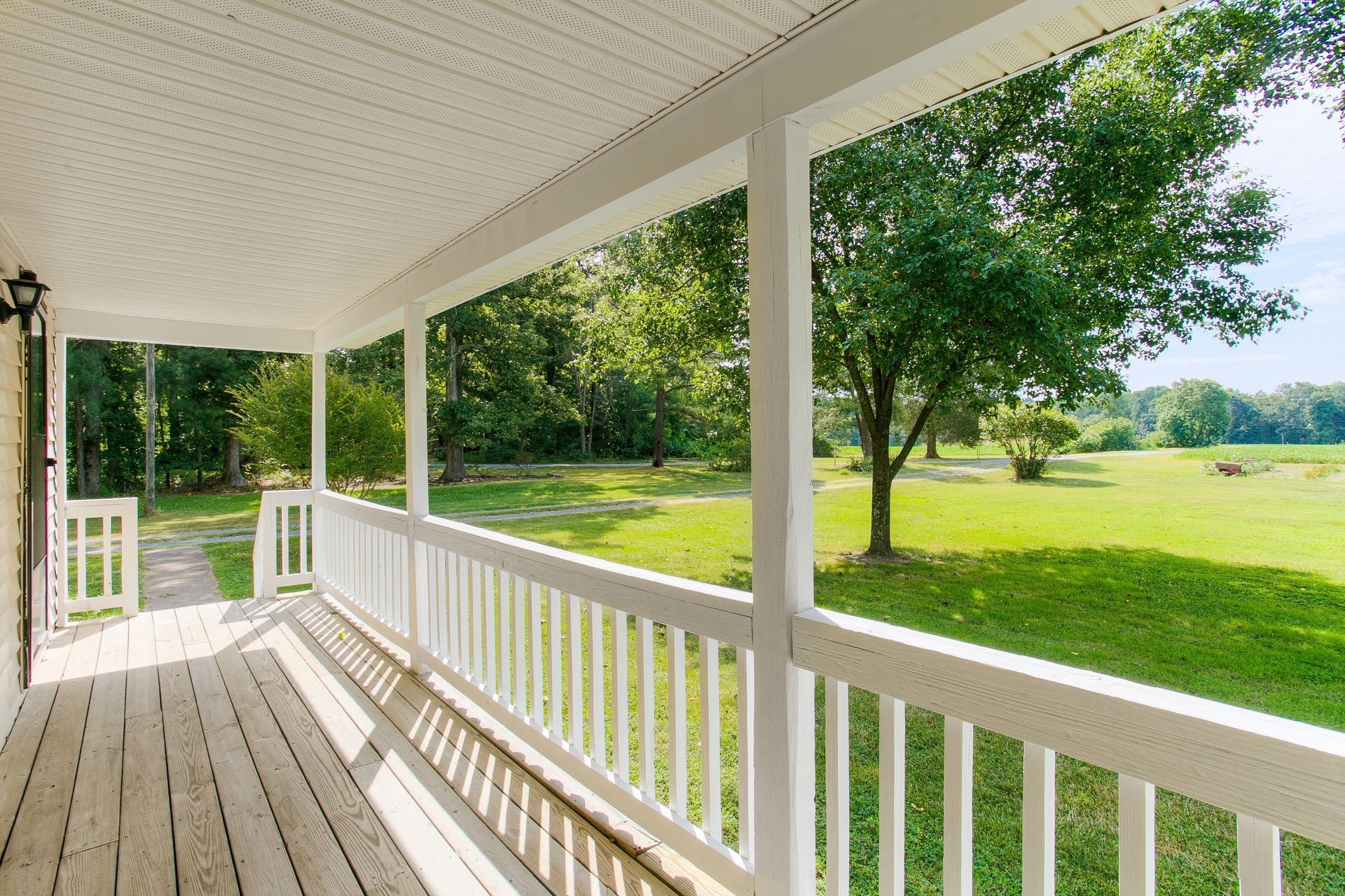 White porch with railings overlooks a green field and trees under a blue sky.