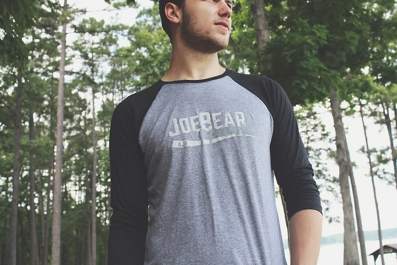 A man wearing a joe bear baseball shirt is standing in the woods.