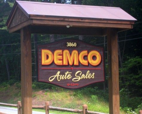Sign for Demco Auto Sales, 3860 address. Wooden post with brown sign, yellow lettering, and a brown roof.