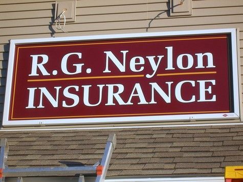 Sign for R.G. Neylon Insurance; white text on a burgundy background, white-framed, mounted above a brown roof.