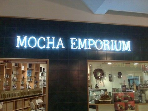Mocha Emporium storefront with illuminated white letters, a dark tiled background, and displays of products.
