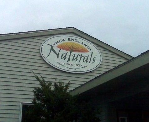 Sign for New England Naturals, an oval sign with a tree logo, on a building with siding.