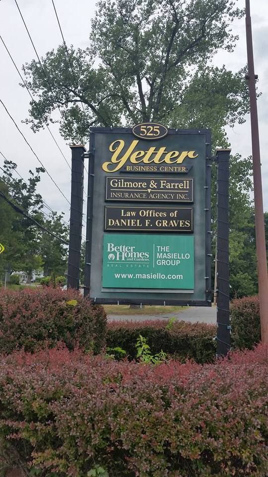 Sign for Yetter Business Center, listing businesses. Green and gold signage against a backdrop of bushes and trees.