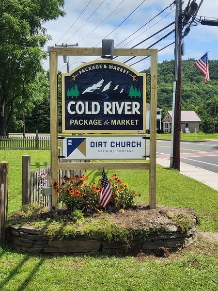 Sign for Cold River Package & Market with American flag, flowers, and Dirt Church banner, in a green, outdoor setting.