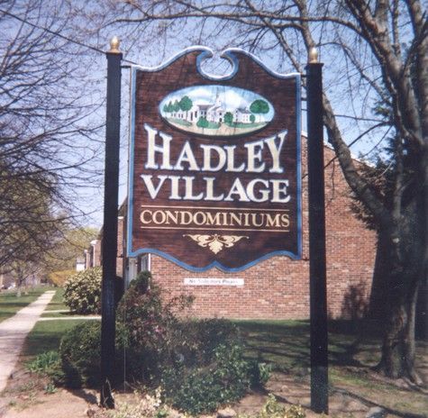 Sign for Hadley Village Condominiums: brown, white text, illustration of a house, brick building background.