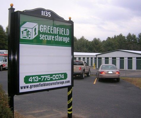 Sign for Greenfield Secure Storage; storage units in background, car parked in front.