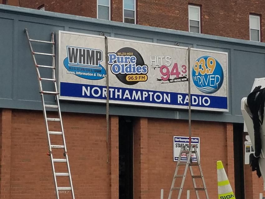 Sign for Northampton Radio with station logos, attached to building. Ladders and cones visible.