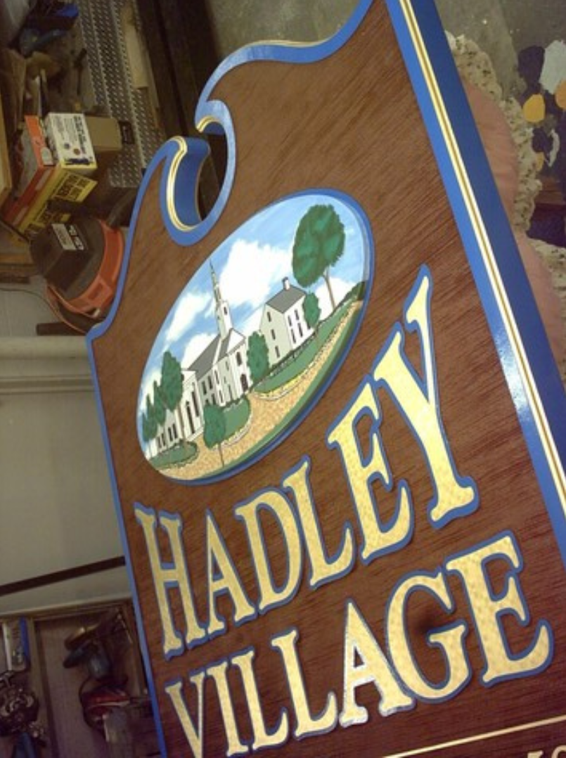 Sign for Hadley Village with illustration of a church, gold lettering, brown wood, and blue and gold trim.