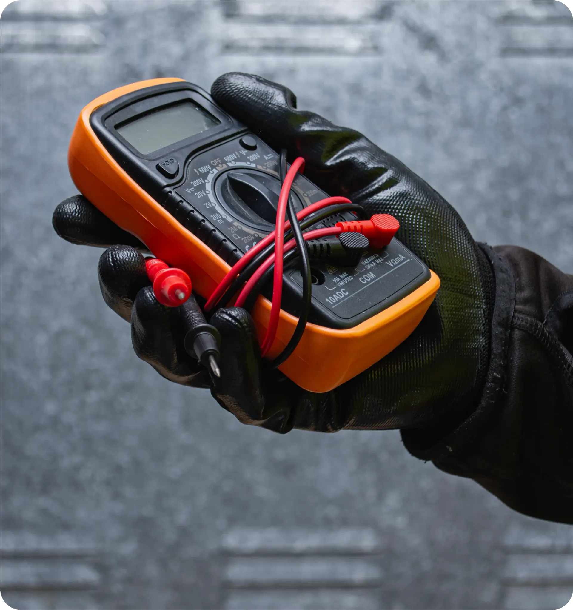 Gloved hand holding an orange and black multimeter with red and black probes, in front of a gray surface.