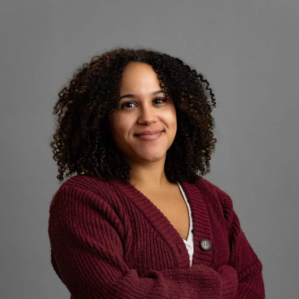 Woman with curly brown hair wearing a burgundy cardigan smiles with arms crossed, against a gray background.