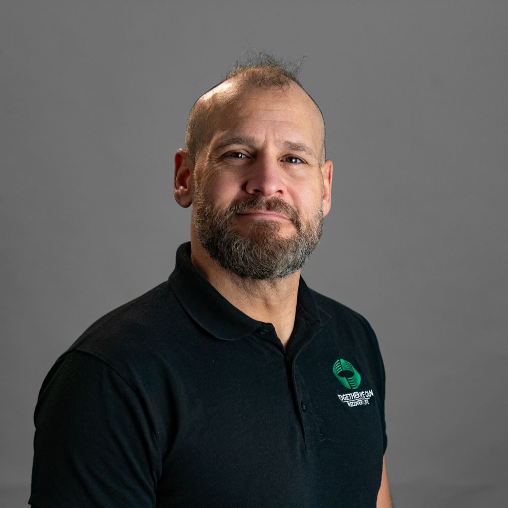 A man in a grey shirt is smiling with his arms crossed.